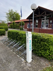 Fahrrad Ladestation am Freibad Hengstforde.jpg Vor einem Backsteingebäude steht eine Fahrrad-Ladestation mit Metallfahrradständern, umgeben von gepflegter Hecke.