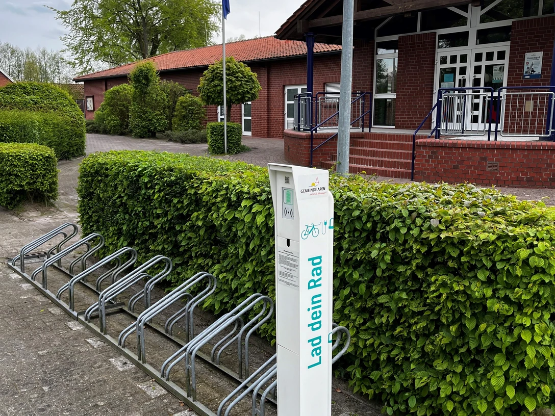 Fahrrad Ladestation am Freibad Hengstforde.jpg Vor einem Backsteingebäude steht eine Fahrrad-Ladestation mit Metallfahrradständern, umgeben von gepflegter Hecke.