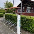 Fahrrad Ladestation am Freibad Hengstforde.jpg Vor einem Backsteingebäude steht eine Fahrrad-Ladestation mit Metallfahrradständern, umgeben von gepflegter Hecke.