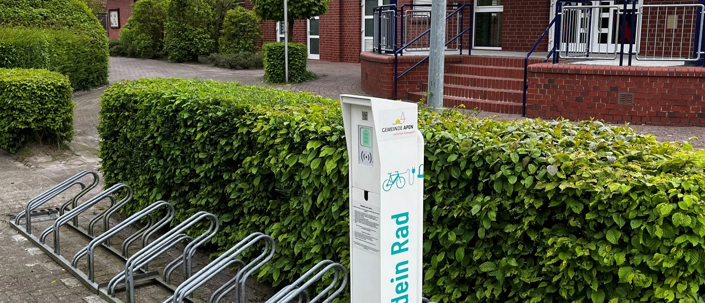 Fahrrad Ladestation am Freibad Hengstforde.jpg Vor einem Backsteingebäude steht eine Fahrrad-Ladestation mit Metallfahrradständern, umgeben von gepflegter Hecke.