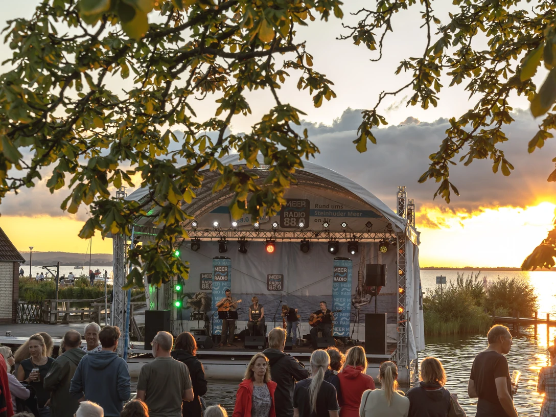 Seebühne 1 Eine Seebühne am Steinhuder Meer, umrahmt von Bäumen, bei Sonnenuntergang mit Publikum und Musikern.