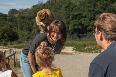 Eine Frau lächelt, während ein Affe auf ihrer Schulter sitzt, umgeben von lachenden Menschen im Freien.