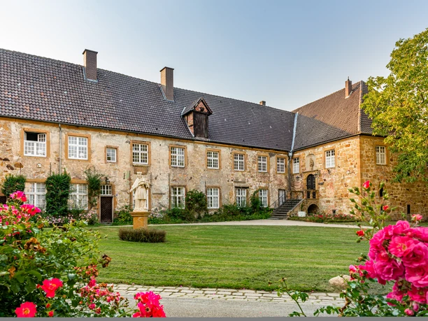 Teutoburger Wald, Klosteranlage Herzebrock, M. Wallenfang Historisches Schloss mit Rosengarten und Statuen, umgeben von grüner Landschaft und blauer Himmel.