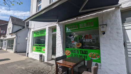Pizza Haus Bad Oeynhausen Zu sehen ist die Fassade des Pizzahauses, die in einem neutralen Weiß gehalten ist. Auf großen Fenstern sind verschiedene Schriftzüge zu sehen, und grüne Farbtöne kommen zum Einsatz. Vorne steht ein Tisch.
