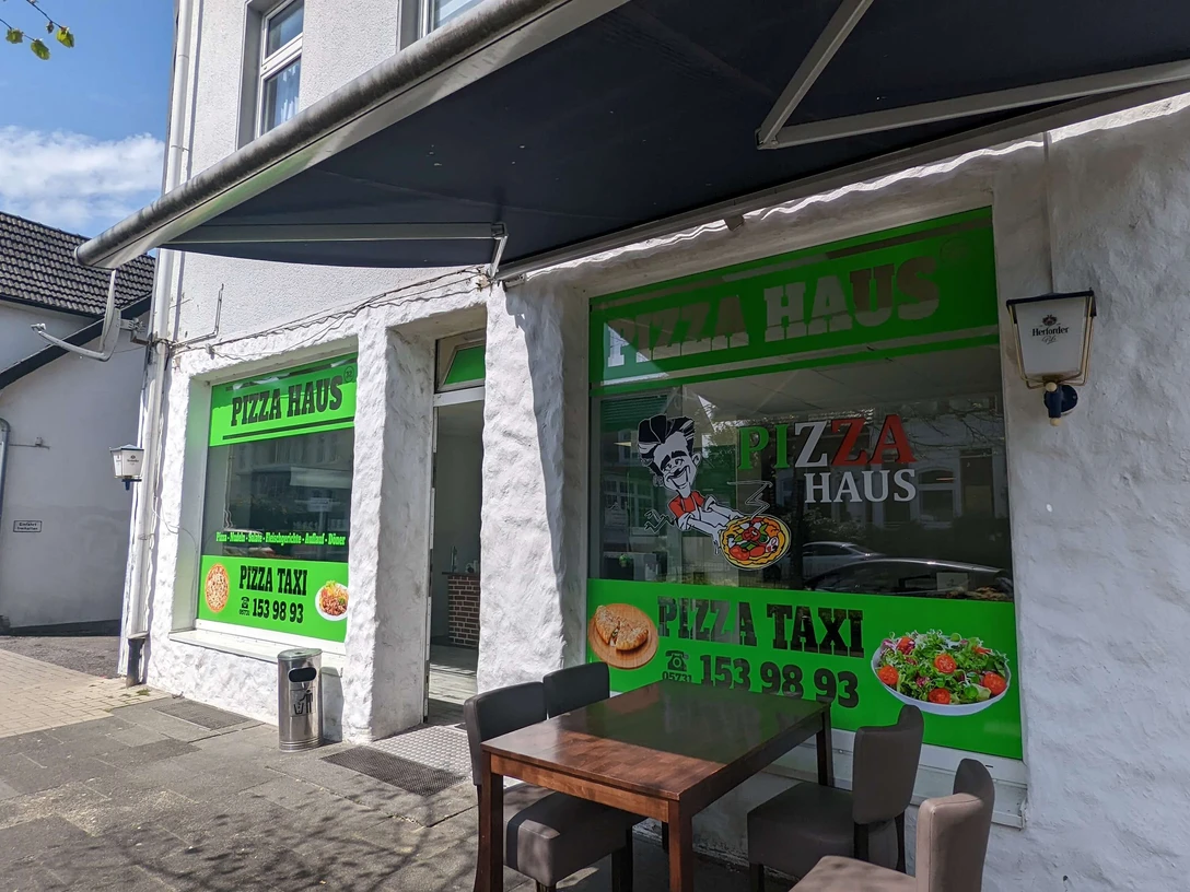 Pizza Haus Bad Oeynhausen Zu sehen ist die Fassade des Pizzahauses, die in einem neutralen Weiß gehalten ist. Auf großen Fenstern sind verschiedene Schriftzüge zu sehen, und grüne Farbtöne kommen zum Einsatz. Vorne steht ein Tisch.