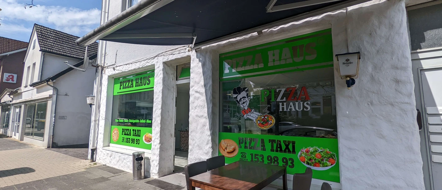 Pizza Haus Bad Oeynhausen Zu sehen ist die Fassade des Pizzahauses, die in einem neutralen Weiß gehalten ist. Auf großen Fenstern sind verschiedene Schriftzüge zu sehen, und grüne Farbtöne kommen zum Einsatz. Vorne steht ein Tisch.