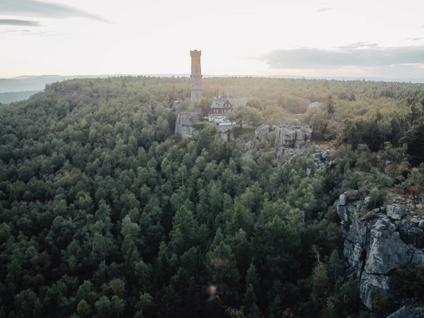 Hoher Schneeberg Ein hoher Turm und ein Gebäude stehen auf einem bewaldeten Hügel, umgeben von dichtem Grün und Felsen, bei sanftem Sonnenlicht.