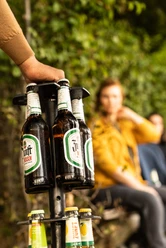 Bierweg Bierflaschen auf einem Flaschenhalter im Vordergrund, unscharfe Personen im Hintergrund.