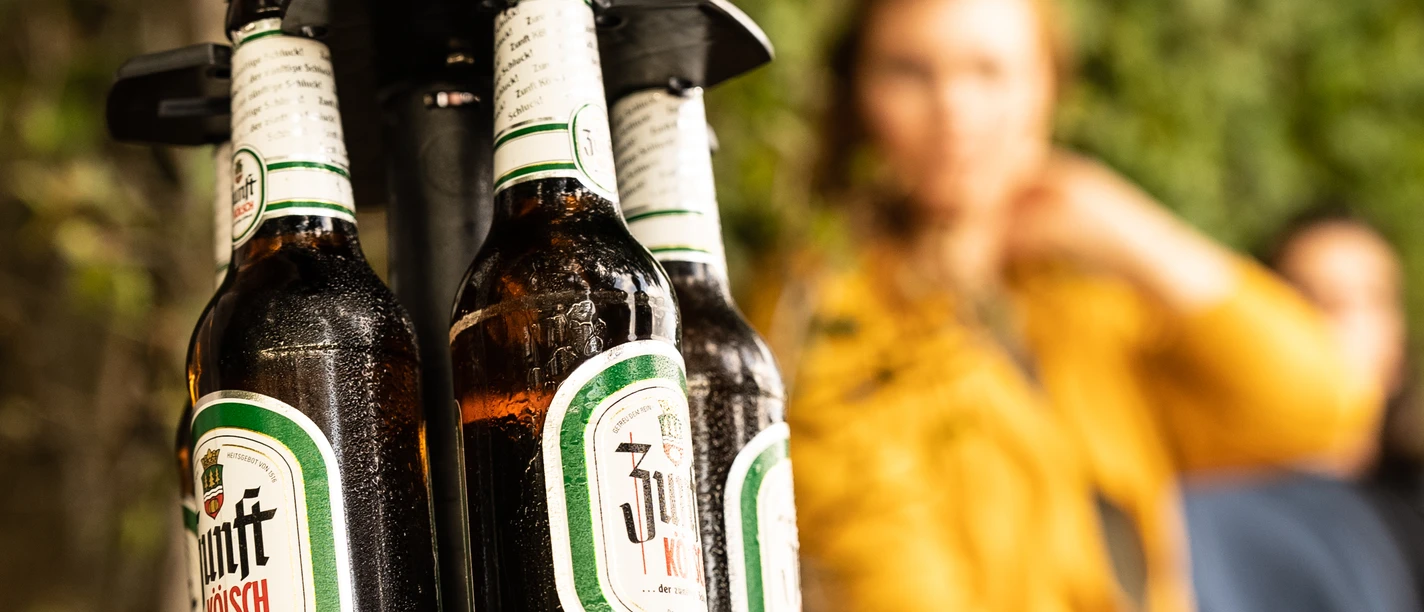 Bierweg Bierflaschen auf einem Flaschenhalter im Vordergrund, unscharfe Personen im Hintergrund.