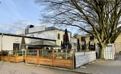 Eine Eventlocation mit großzügiger Außenterrasse, geschmückt mit Sonnenschirmen und umgeben von Bäumen.