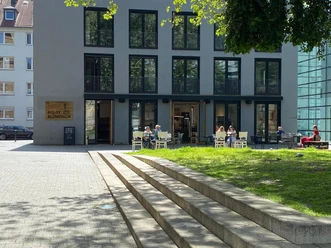 Ein Außenbereich eines Cafés mit Gästen vor einem modernen, mehrstöckigen Gebäude bei sonnigem Wetter.
