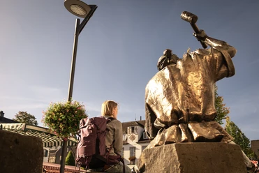 Altstadt Wipperfürth Ein Tourist sitzt neben einer Bronzestatue auf einem Platz unter blauem Himmel in Wipperfürth.