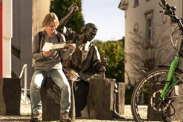 Altstadt Wipperfürth Junger Mann studiert eine Karte neben einer Bronzestatue auf Steinblöcken in sonniger Straßenumgebung.