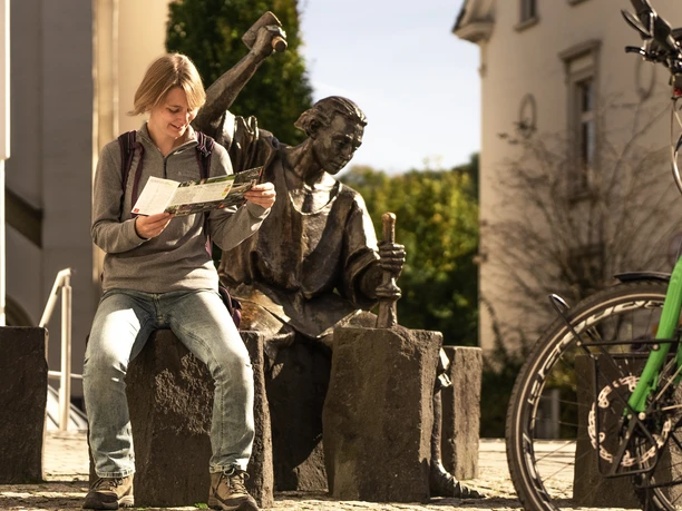 Altstadt Wipperfürth Junger Mann studiert eine Karte neben einer Bronzestatue auf Steinblöcken in sonniger Straßenumgebung.