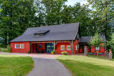 Teutoburger Wald / Museum Peter August Böckstiegel / Werther (Westf.) / Foto: M. Wallenfang Farbkräftiges rotes Haus im Fachwerkstil mit blau umrahmter Tür, umgeben von üppigem Grün.