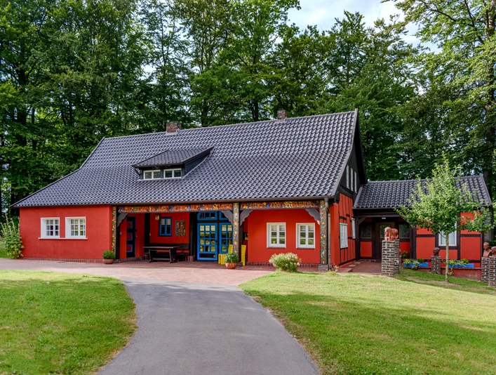 Teutoburger Wald / Museum Peter August Böckstiegel / Werther (Westf.) / Foto: M. Wallenfang Farbkräftiges rotes Haus im Fachwerkstil mit blau umrahmter Tür, umgeben von üppigem Grün.