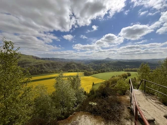 Gezicht op een weids landschap met gele koolzaadvelden, groene weiden en heuvels onder een blauwe wolkenlucht, een reling op de voorgrond.