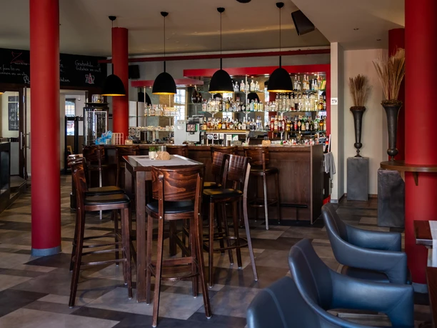 Zinnober-37.jpg Een stijlvol ingericht restaurant met hoge rode zuilen, houten meubels en een verlichte bar.