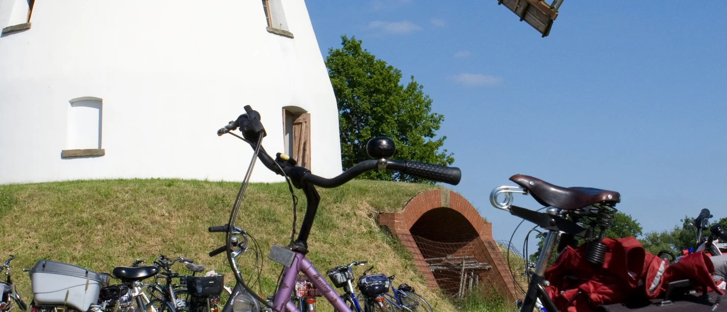Stadt Petershagen_Heimsen_Evelyn Hotze_Mühle_außen_einzelnes Rad im Vordergrund_Sommer_hoch.jpg Historische Windmühle mit langem Flügelwerk in ruhigem Umfeld; Fahrrad im Vordergrund.