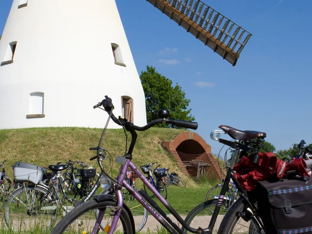 Stadt Petershagen_Heimsen_Evelyn Hotze_Mühle_außen_einzelnes Rad im Vordergrund_Sommer_hoch.jpg Historische Windmühle mit langem Flügelwerk in ruhigem Umfeld; Fahrrad im Vordergrund.