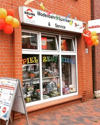 Ansicht.png Ein rotes Backsteingebäude mit einem Schaufenster voller Spielzeug und bunten Luftballons vorne.