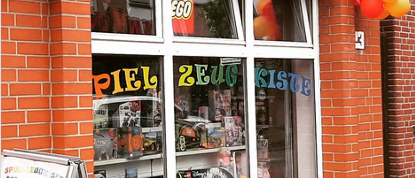 Ansicht.png Ein rotes Backsteingebäude mit einem Schaufenster voller Spielzeug und bunten Luftballons vorne.