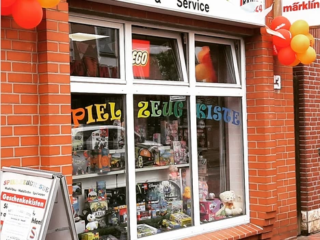 Ansicht.png Ein rotes Backsteingebäude mit einem Schaufenster voller Spielzeug und bunten Luftballons vorne.