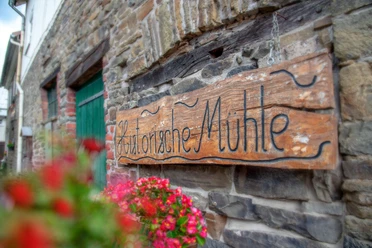 Mühle Nespen Holztafel mit der Aufschrift "Historische Mühle" an einer rustikalen Steinwand, umrahmt von roten Blumen.