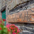 Mühle Nespen Holztafel mit der Aufschrift "Historische Mühle" an einer rustikalen Steinwand, umrahmt von roten Blumen.