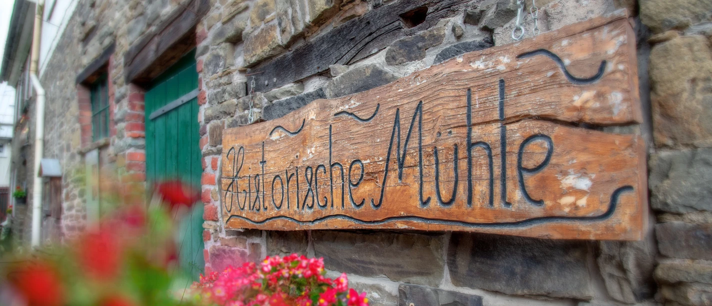 Mühle Nespen Holztafel mit der Aufschrift "Historische Mühle" an einer rustikalen Steinwand, umrahmt von roten Blumen.