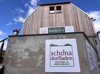 Schöna Dorfladen Fassade eines Gebäudes mit Holzverkleidung, Schildern des "Schöna Dorfladen", blauer Himmel, links eine Gießkanne mit Blumen.