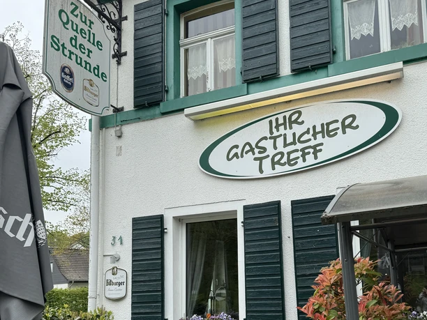 Zur Quelle der Strunde - Außenansicht Gemütliches Gasthaus "Zur Quelle der Strunde" mit grünen Fensterläden und einladendem Eingang.