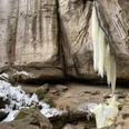 Gautschgrotte im Winter Gefrorener Wasserfall hängt von einer steinigen Klippe, umgeben von schneebedeckten Bäumen und Felsen in einer winterlichen Landschaft.