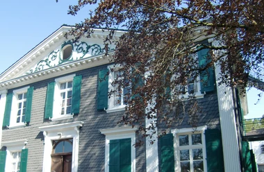 Pulvermuseum Villa Ohl Zweistöckiges historisches Gebäude mit grünen Fensterläden, verziert mit Stuckornamenten und Baum im Vordergrund.