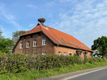 ÖSSM Winzlar Backsteinhaus mit rotem Dach in ländlicher Umgebung, auf dem Dach ein Storchennest unter blauem Himmel.