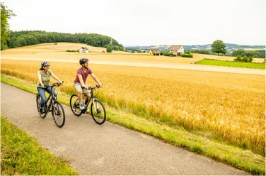 Stadtrand.jpg Zwei Radfahrer auf einem asphaltierten Weg durch eine goldene Agrarlandschaft bei bewölktem Himmel.