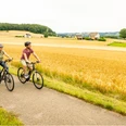 Stadtrand.jpg Zwei Radfahrer auf einem asphaltierten Weg durch eine goldene Agrarlandschaft bei bewölktem Himmel.