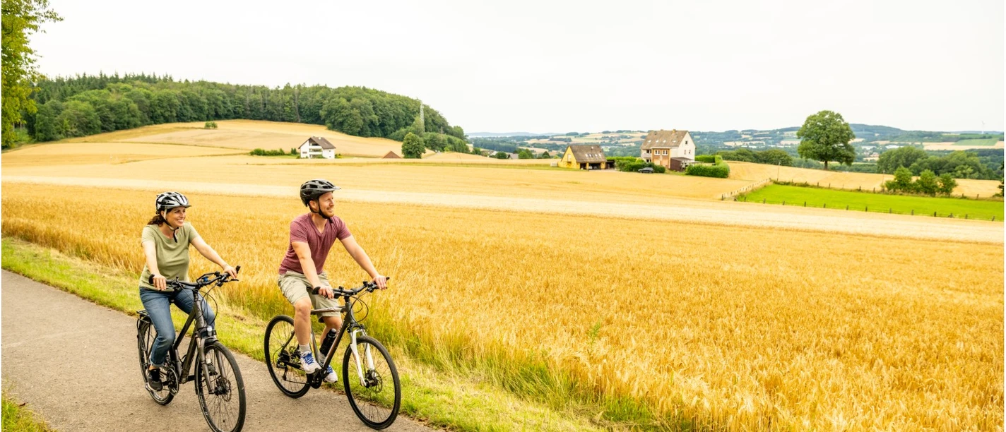 Stadtrand.jpg Zwei Radfahrer auf einem asphaltierten Weg durch eine goldene Agrarlandschaft bei bewölktem Himmel.