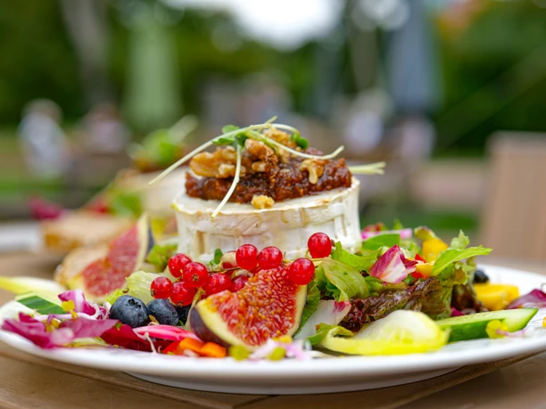 Ein voller Teller in sommerlicher Umgebung. Ein großer Salat aus Gemüse und Obst mit einem überbackenen Käse.