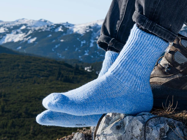 steppenstrolch 8 Socken aus Schafwolle Füße, in blauen Socken aus Schafswolle gehüllt, stützen sich auf einen Fels. Im Hintergrund sind verschneite Berge.