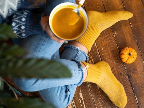 steppenstrolch 9 Socken aus Schafwolle Person in heller Jeans und gelben Socken, trinkt Suppe auf einem Holzboden, ein Kürbis liegt daneben.