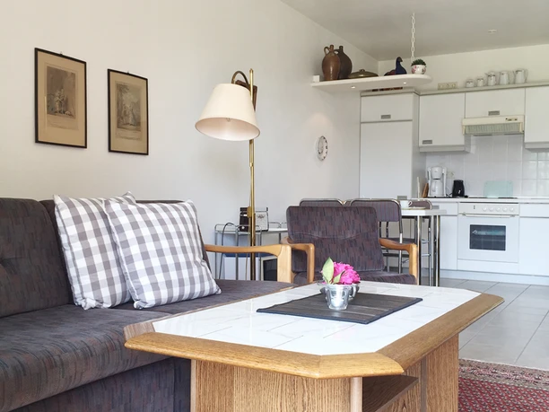 gemütlicher Wohnbereich mit Zugang zur Terrasse Wohnbereich mit Sofa, karierten Kissen, Kacheltisch und offener weißer Küche.
