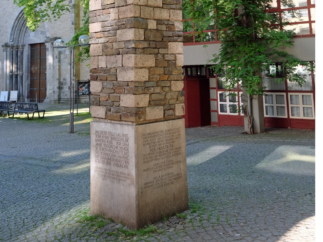 Schmitz column Ein hoher, mit unterschiedlich farbigen Steinen verkleideter Pfeiler steht auf einem gepflasterten Platz vor einer Kirche. Umgeben von Bäumen und einem Gebäude mit roten Fensterrahmen.A tall pillar clad with differently colored stones stands on a paved square in front of a church. It is surrounded by trees and a building with red window frames.