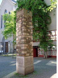 Schmitz Säule Ein hoher, mit unterschiedlich farbigen Steinen verkleideter Pfeiler steht auf einem gepflasterten Platz vor einer Kirche. Umgeben von Bäumen und einem Gebäude mit roten Fensterrahmen.