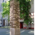 Schmitz Säule Ein hoher, mit unterschiedlich farbigen Steinen verkleideter Pfeiler steht auf einem gepflasterten Platz vor einer Kirche. Umgeben von Bäumen und einem Gebäude mit roten Fensterrahmen.A tall pillar clad with differently colored stones stands on a paved square in front of a church. It is surrounded by trees and a building with red window frames.