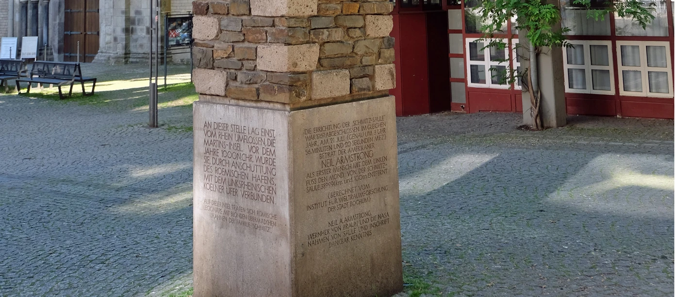 Schmitz Säule Ein hoher, mit unterschiedlich farbigen Steinen verkleideter Pfeiler steht auf einem gepflasterten Platz vor einer Kirche. Umgeben von Bäumen und einem Gebäude mit roten Fensterrahmen.
