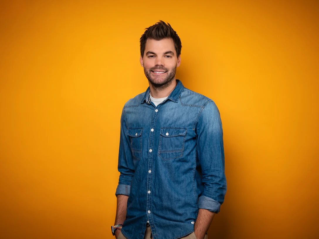 Ruven Rintelmann A man in a denim shirt against a bright orange background smiles at the camera.