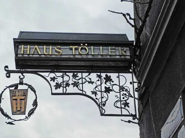 Haus Töller Ein kunstvoll geschmiedetes Schild mit "Haus Töller" hängt an einer Hauswand aus grauem Putz.