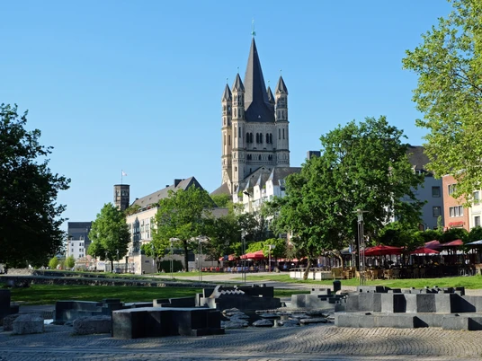 Paolozzibrunnen Im Vordergrund des Bildes befinden sich moderne Kunstwerke aus dunklem Stein, die harmonisch mit der üppigen Begrünung des Parks verschmelzen. Im Hintergrund hebt sich der imposante Turm der Kirche klar gegen den strahlend blauen Himmel ab, flankiert von stilvollen Stadthäusern.In the foreground of the picture are modern works of art made of dark stone, which blend harmoniously with the lush greenery of the park. In the background, the imposing tower of the church stands out clearly against the bright blue sky, flanked by stylish townhouses.