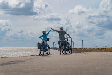 Emden-Außenhafen_01-CC_BY_SA-MaximilianSemsch_EmsRadweg.jpg Zwei Radfahrer am Hafen heben die Hände zum High-Five, im Hintergrund erstreckt sich das Meer.
